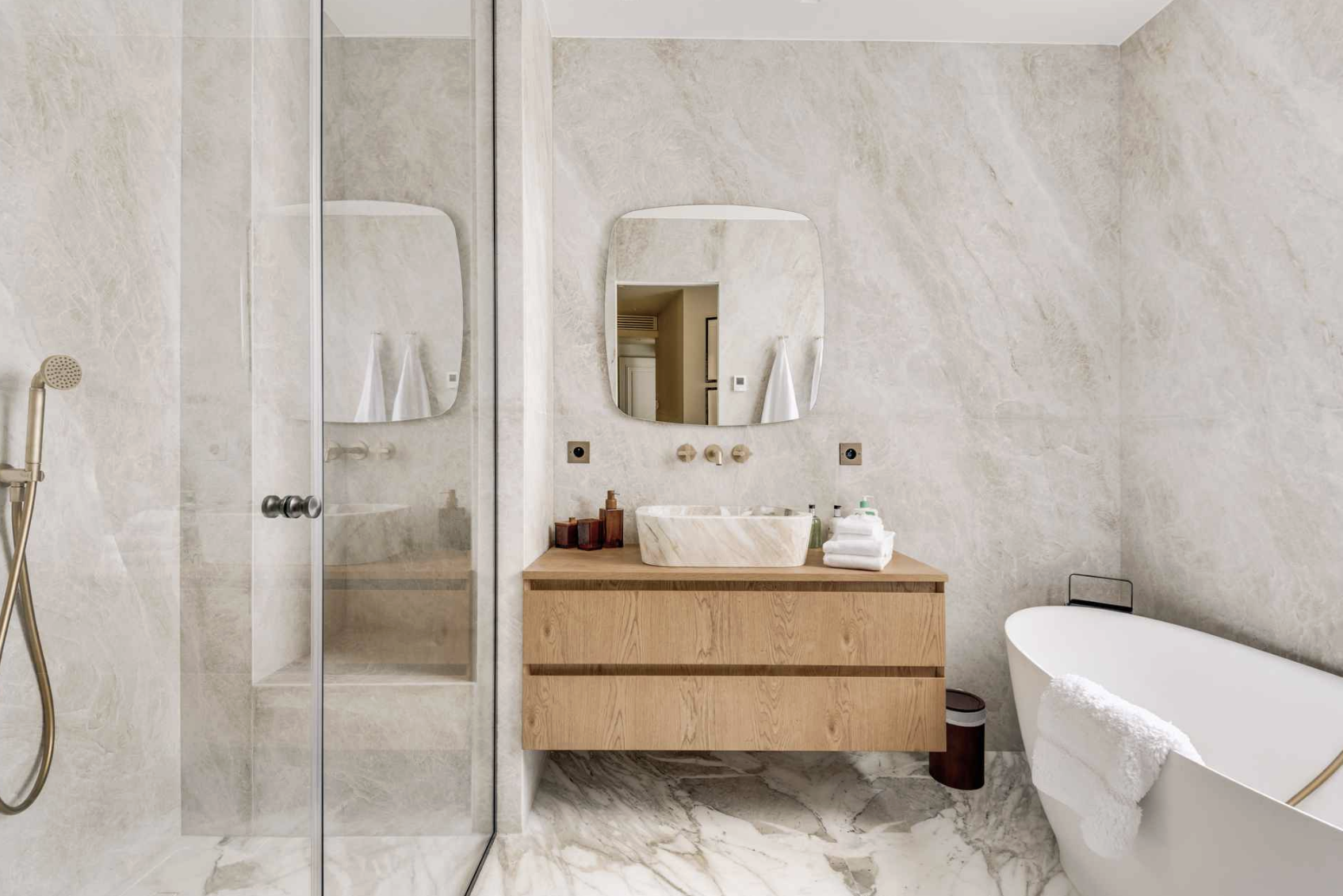 Modern bathroom with marble flooring and walls, glass shower enclosure on the left, wooden vanity with vessel sink, oval mirror, and a freestanding bathtub with a towel.