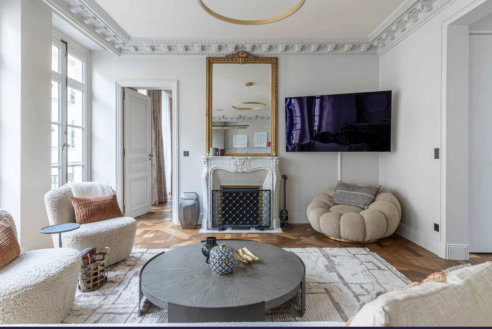 Living room with window, ornate white fireplace, large mirror, wall-mounted TV, textured beige chair, round beige armchair, round coffee table, decorative vases, and hardwood floors.