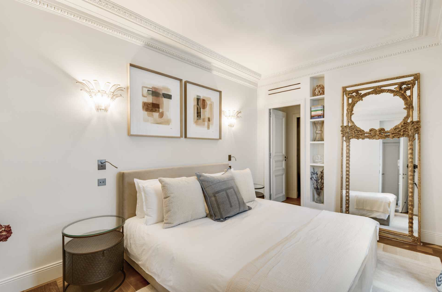 A neatly made bed with multiple decorative pillows in a bedroom with elegant decor. The wall above the bed has two abstract paintings and wall-mounted light fixtures. To the right, there is a large ornate gold-framed mirror and built-in bookshelf wit