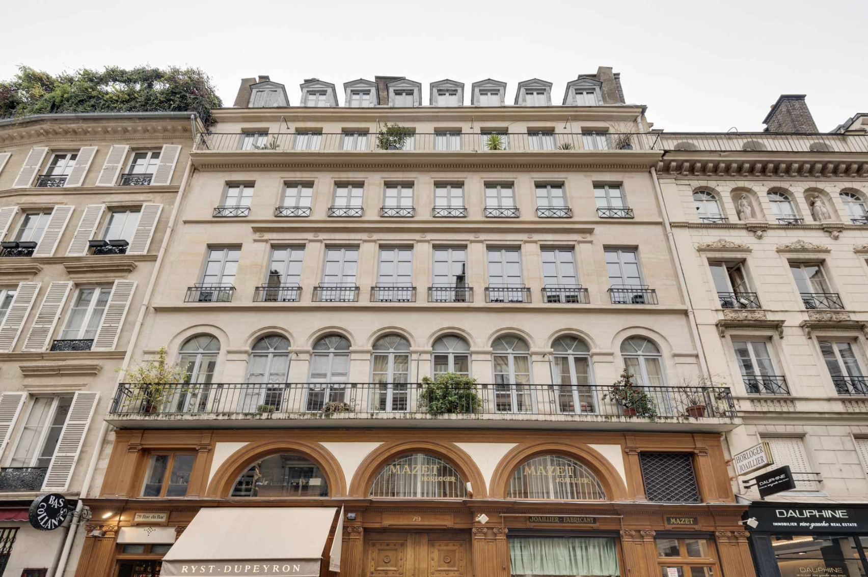 Multi-story Parisian building with retail stores on the ground floor and residential apartments above, with a rooftop garden. The ground floor features storefronts with large arched windows and signage.