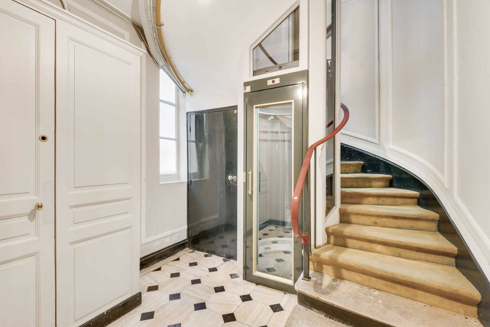 Interior view of a building lobby showing an elevator, staircase, and patterned tile floor with white walls and windows.