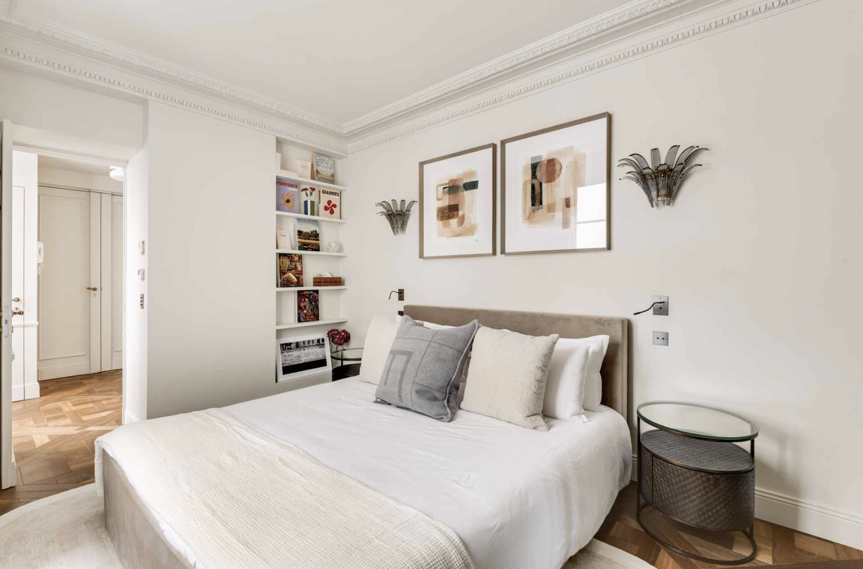 Modern bedroom with white walls, a large bed with white and gray pillows, abstract artwork above, and a small side table with a glass top on the right. Built-in shelves with books and decor are on the left, with parquet hardwood flooring.