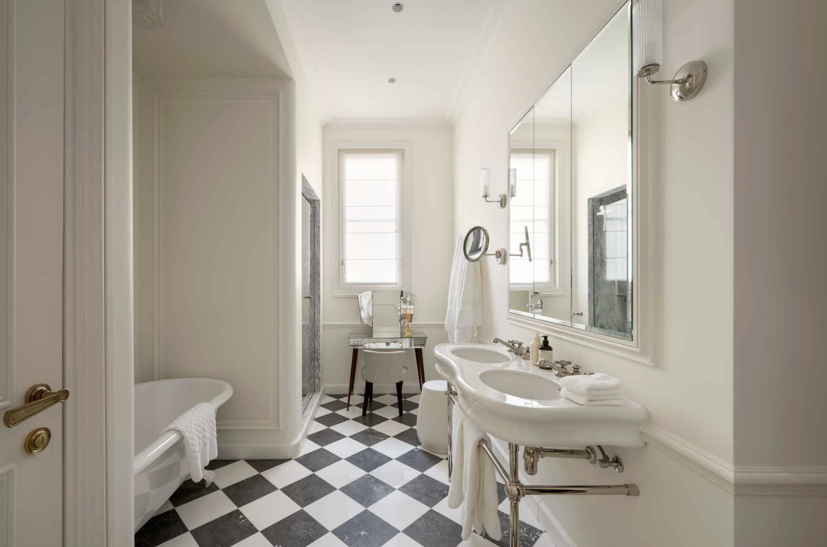 Elegant bathroom with black and white checkered floor, white walls, double sink with large mirror, separate shower with marble walls, clawfoot bathtub with towel, and a small vanity with mirror