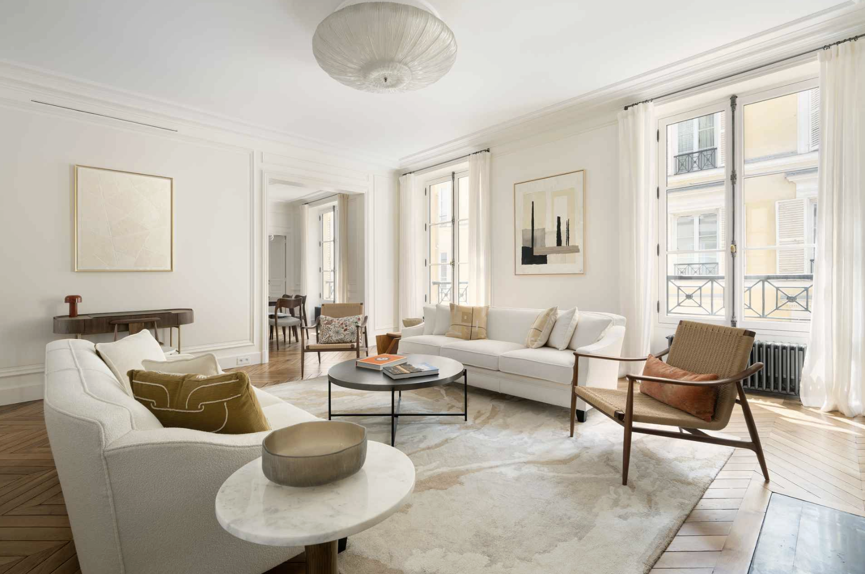 Bright living room with white walls, large windows, white curtains, and minimalist furniture including a white sofa, a beige armchair, and a round coffee table, with artwork on the walls and hardwood floors.