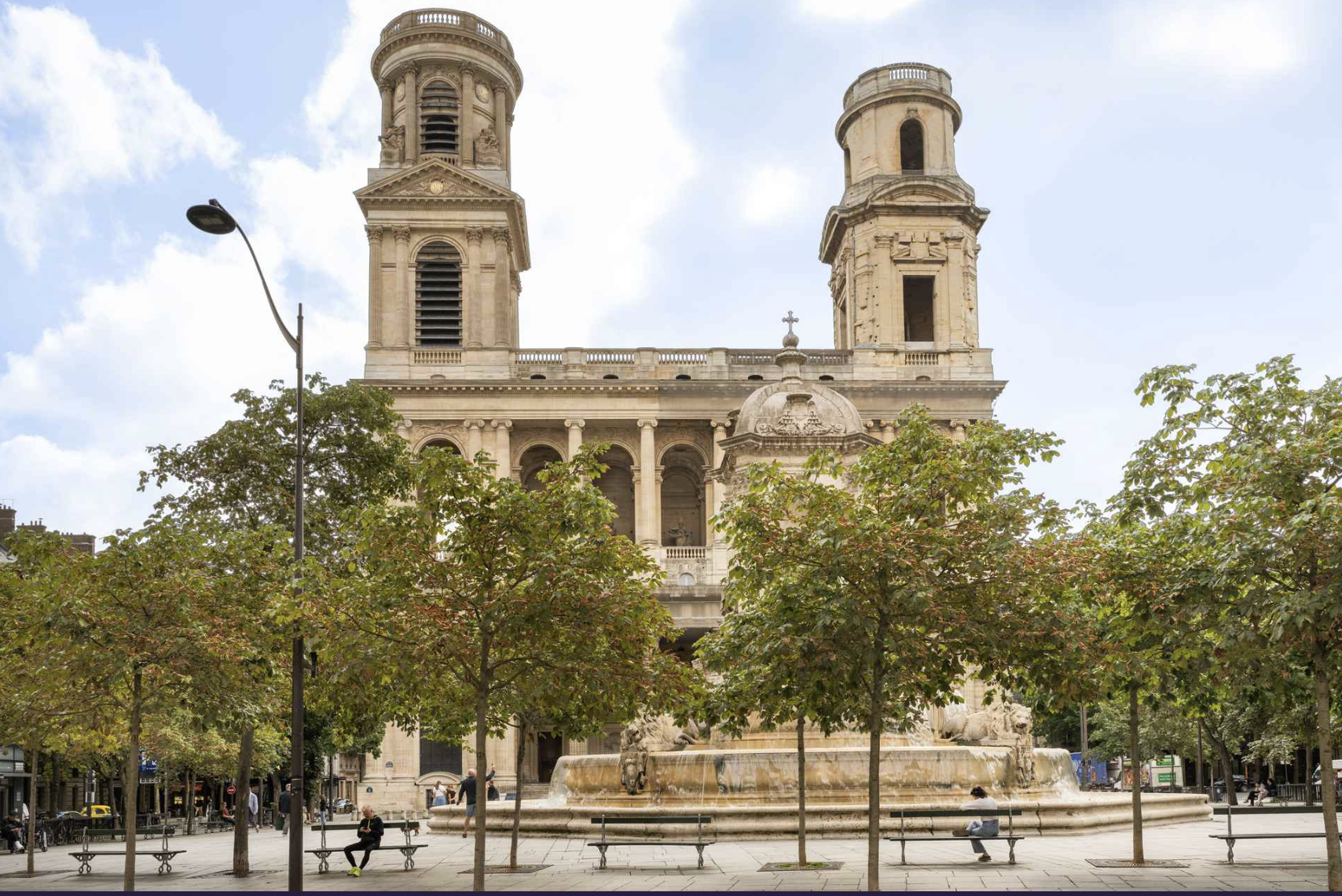 A historic church with two bell towers, situated behind a fountain in a city square. The scene includes daytime with a partly cloudy sky, trees, benches, and a few people sitting and walking around.