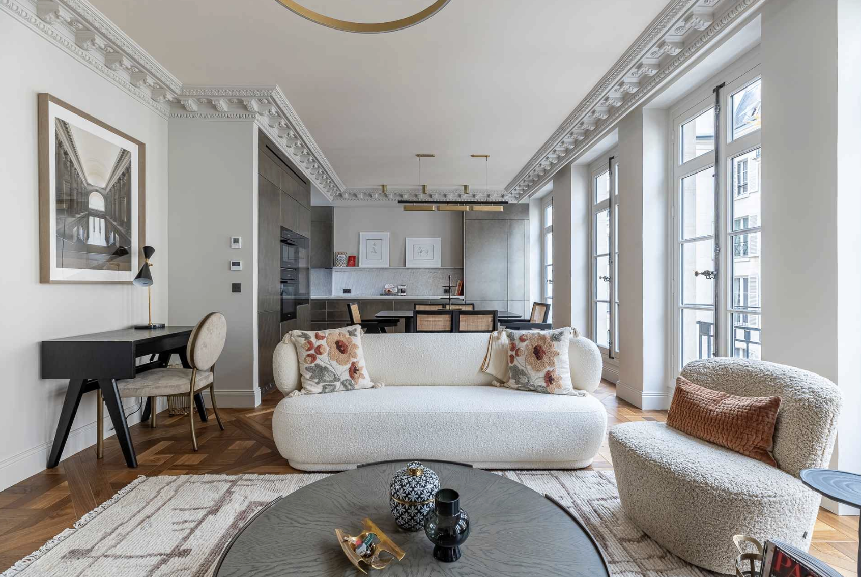 Living room with white sofa, beige armchair, coffee table with decorative items, large windows, and an open kitchen in the background.