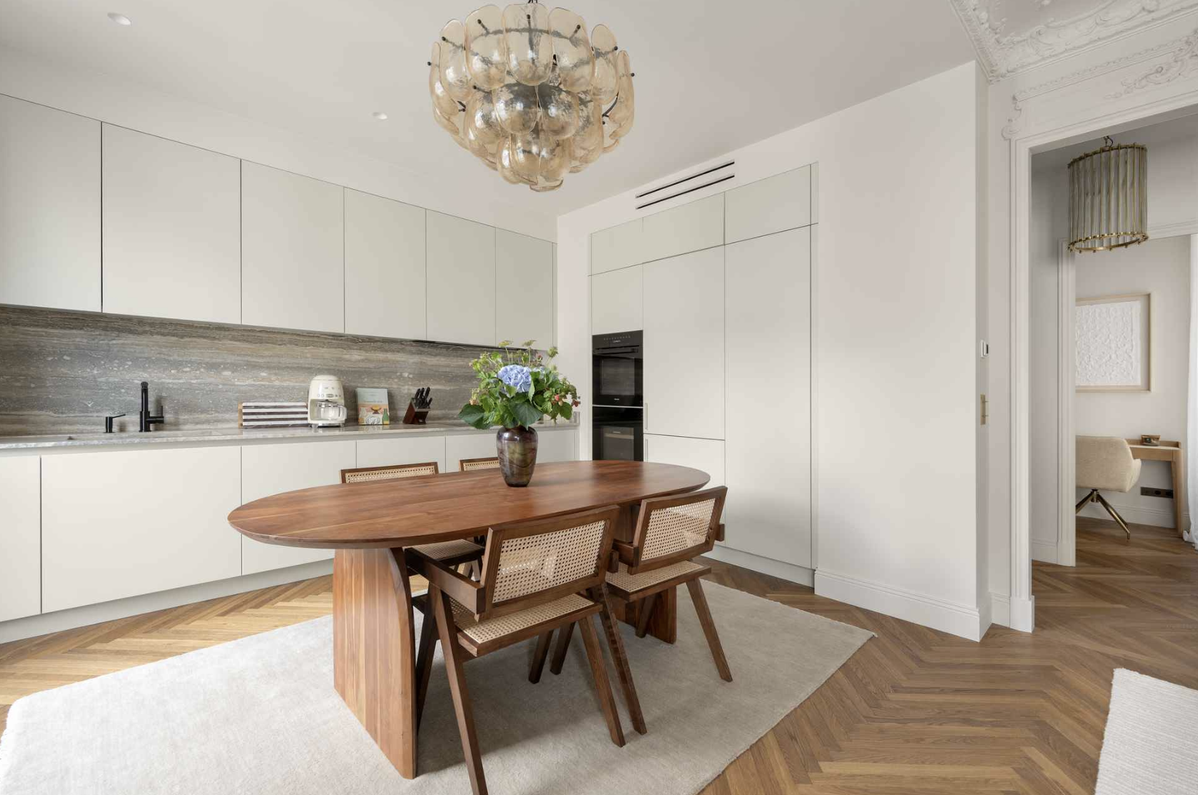 Modern kitchen and dining area with white cabinets, wooden table, four chairs, floral centerpiece, and decorative chandelier.