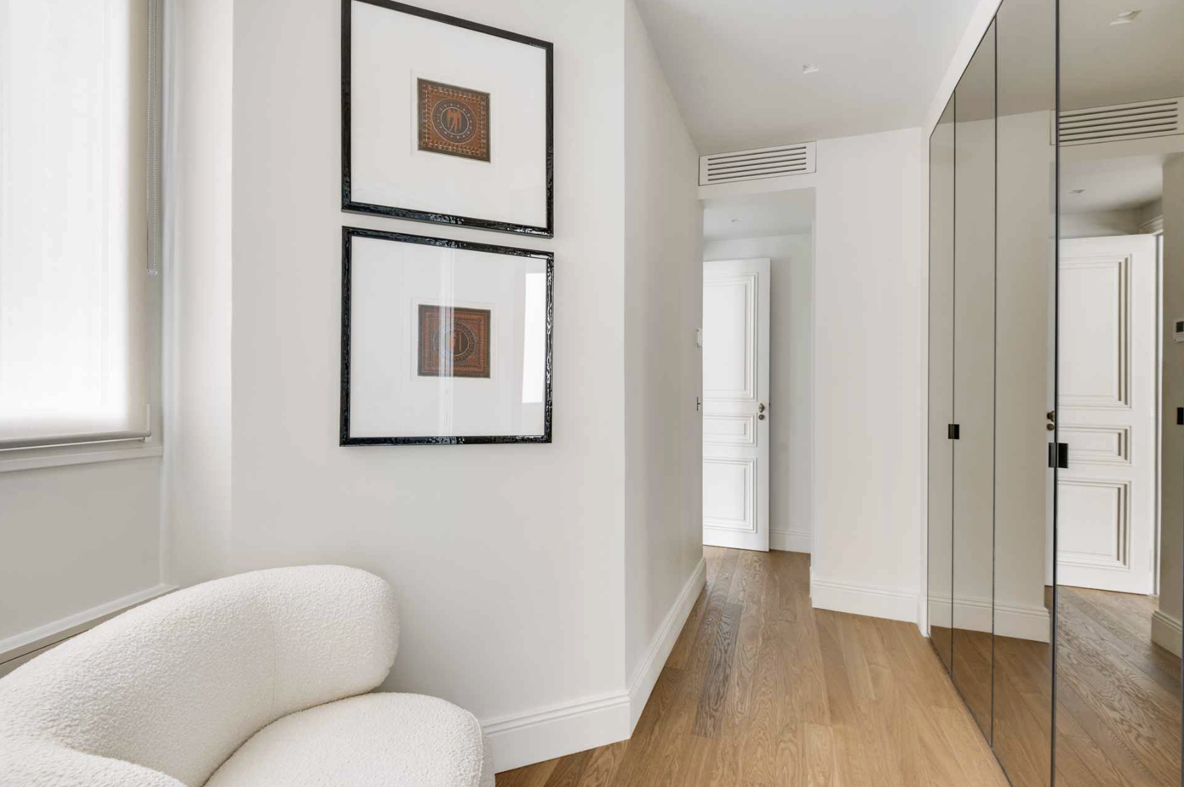 A white textured sofa in a room with white walls and wooden flooring. Two framed artworks hang on the wall above the sofa. There is a window with a white shade on the left. On the right, there is a mirrored closet door and an open white door leading