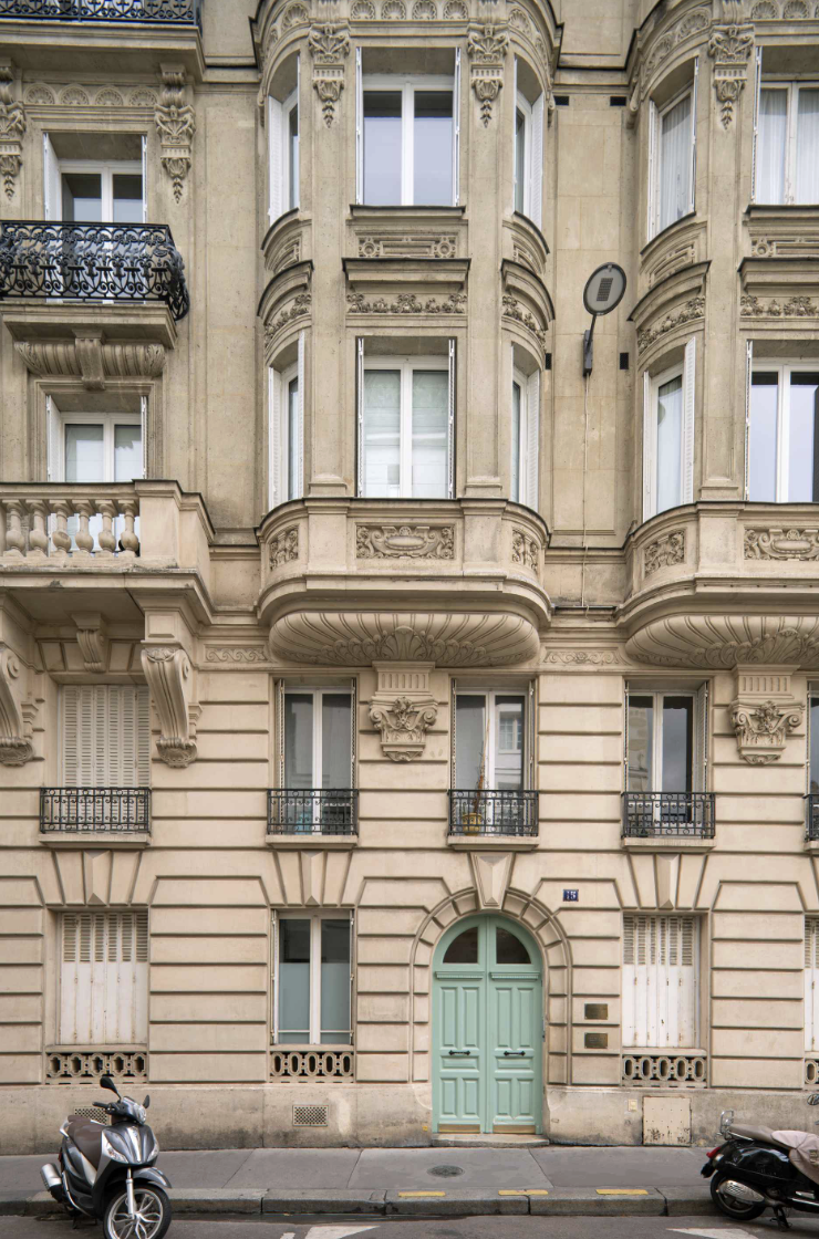 Facade of an ornate, classical-style apartment building with a green arched door, several windows with black and white shutters, decorative stone carvings, small balconies, and two scooters parked in front.
