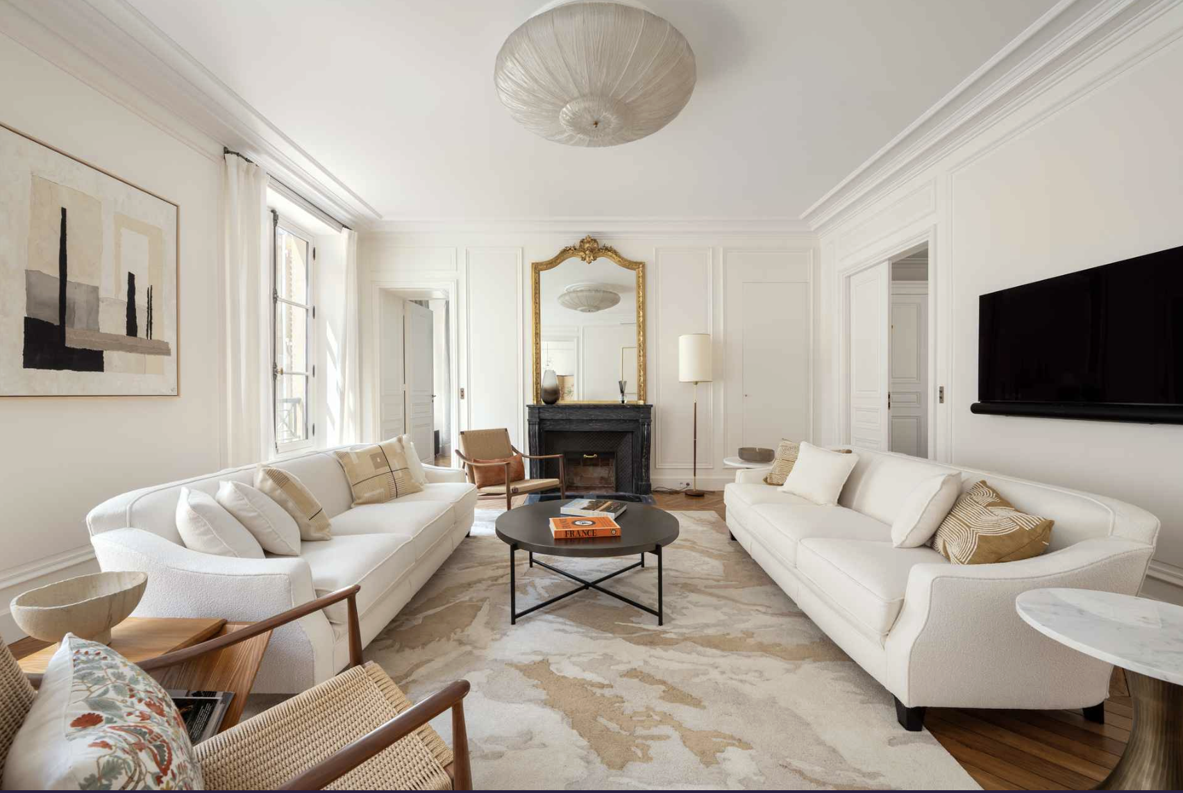 Living room with two white sofas, a black coffee table with books, a fireplace with an ornate mirror, abstract art on the wall, a floor lamp, a wooden chair, and a patterned rug.