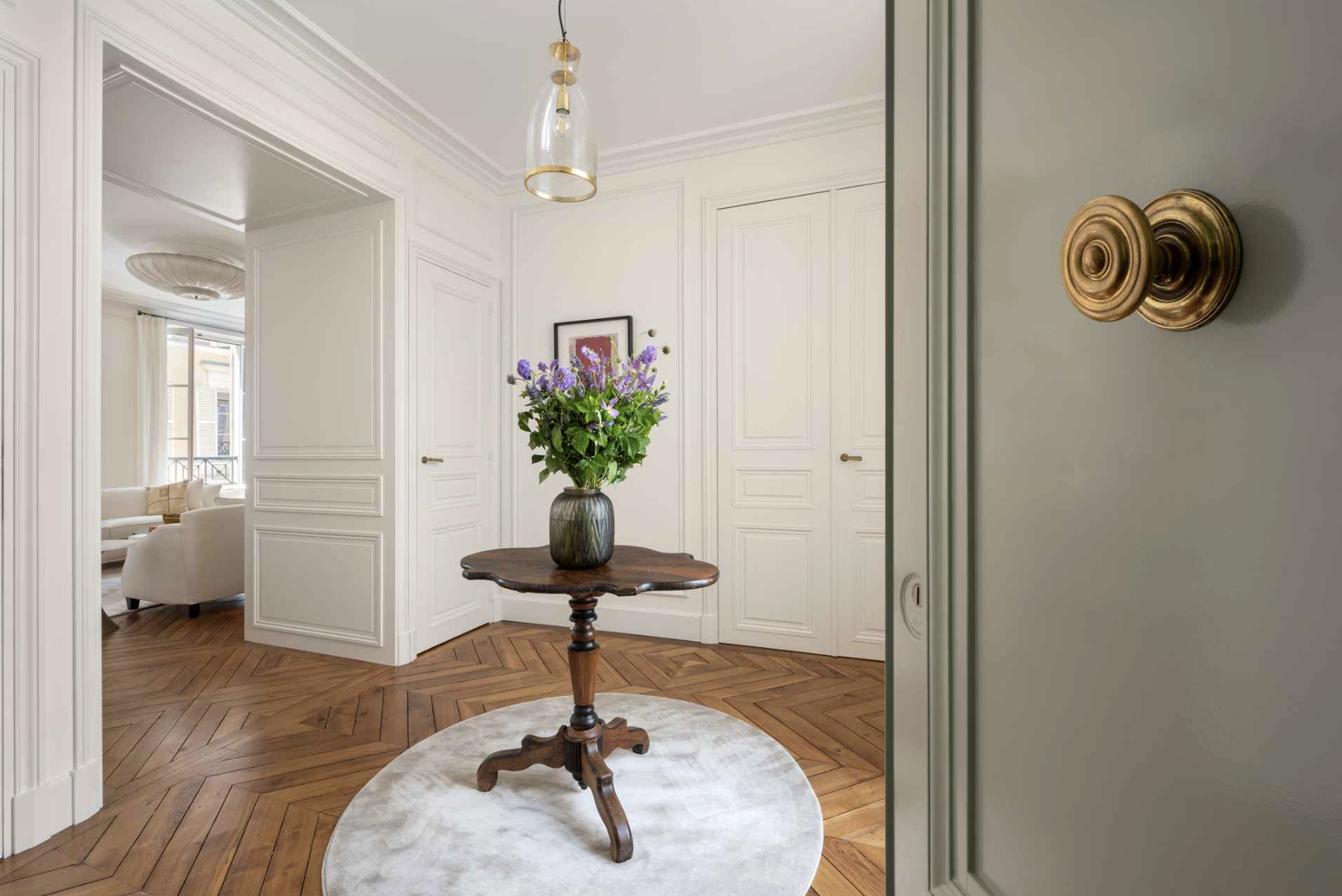Interior of a home with a round wooden table holding a vase of purple flowers, located on a circular rug in a white-walled entryway with polished wood flooring and sliding closet doors.