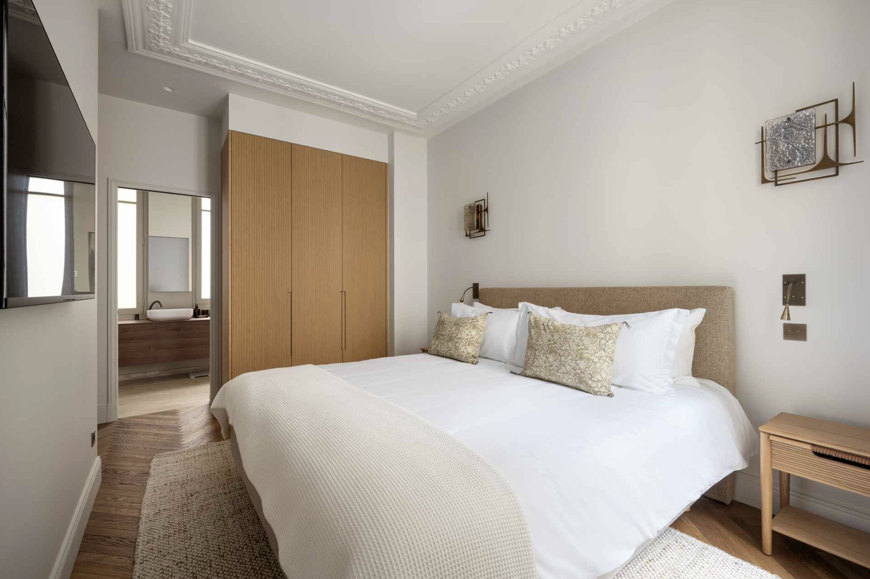 A modern bedroom with a large bed, beige upholstered headboard, white bedding, decorative pillows, wooden side table, and artistic wall sconces. A doorway reveals a bathroom with a bathtub and a window.