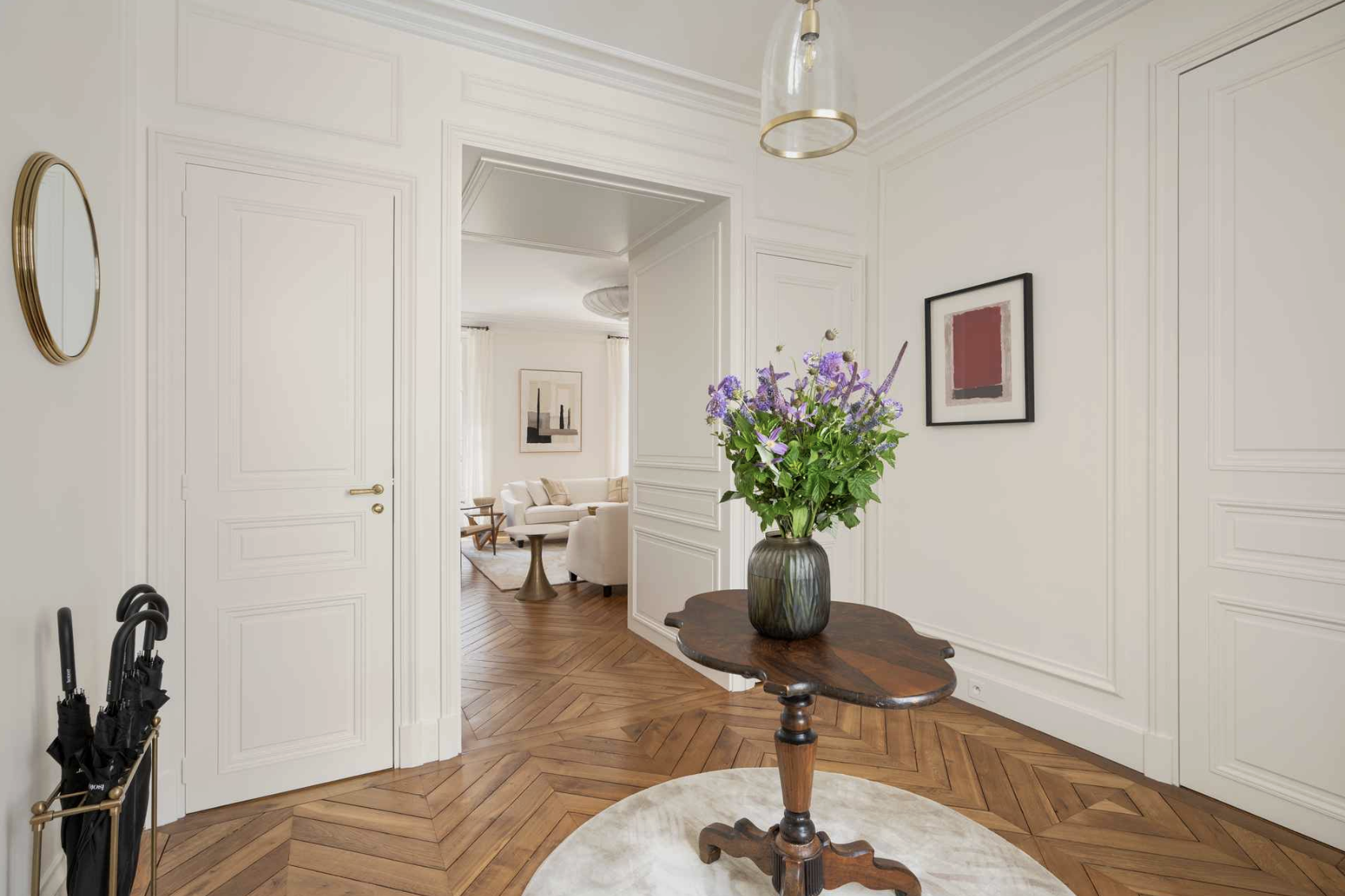 Entryway with wooden floor, white walls, and a wooden table with a purple floral arrangement. Visible doorway leads to a living room with white furniture and art on the walls.