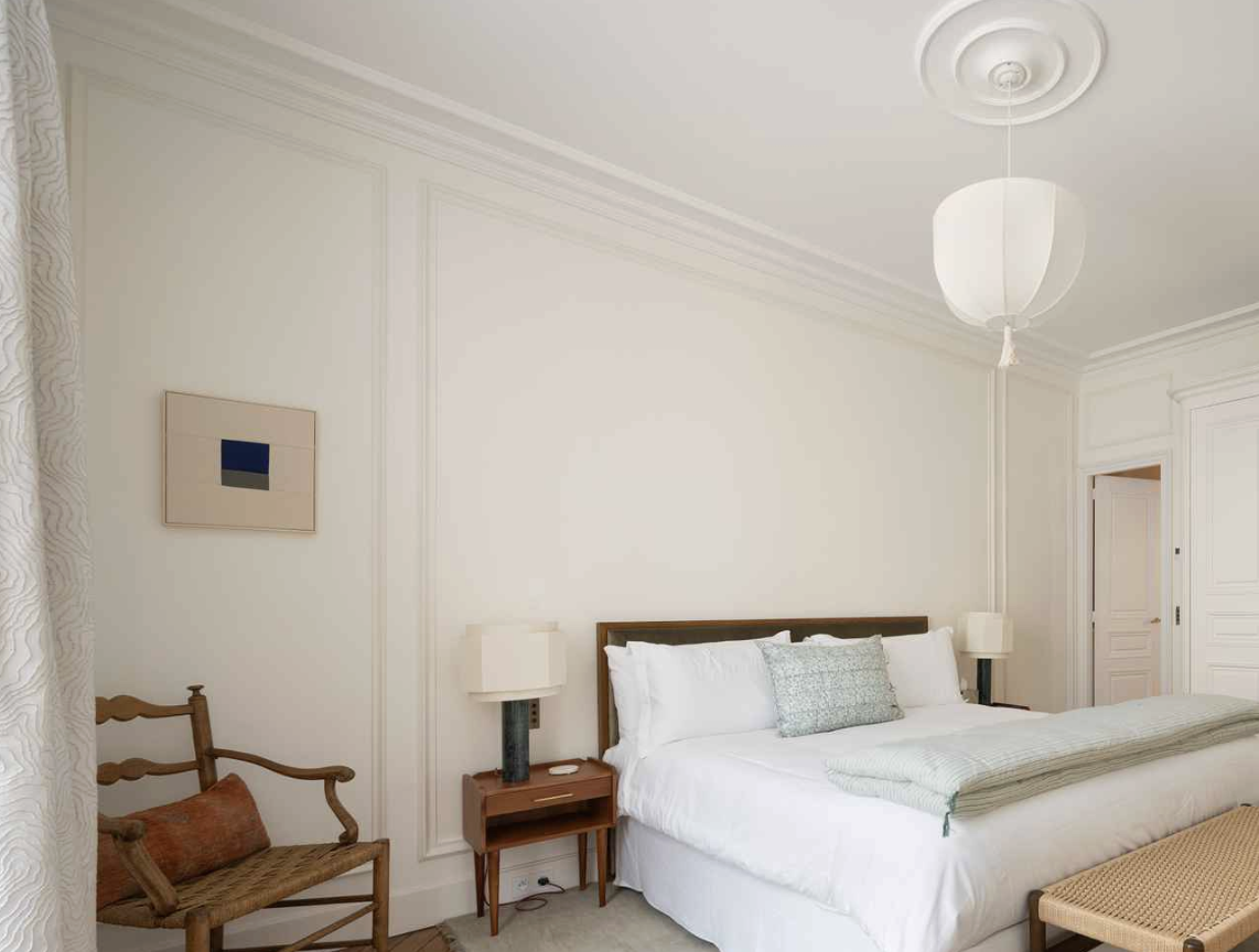 Elegant bedroom with white walls, a bed with white linens and decorative pillows, nightstands with lamps, a wooden armchair, a small framed abstract art piece, and a round ceiling light fixture.