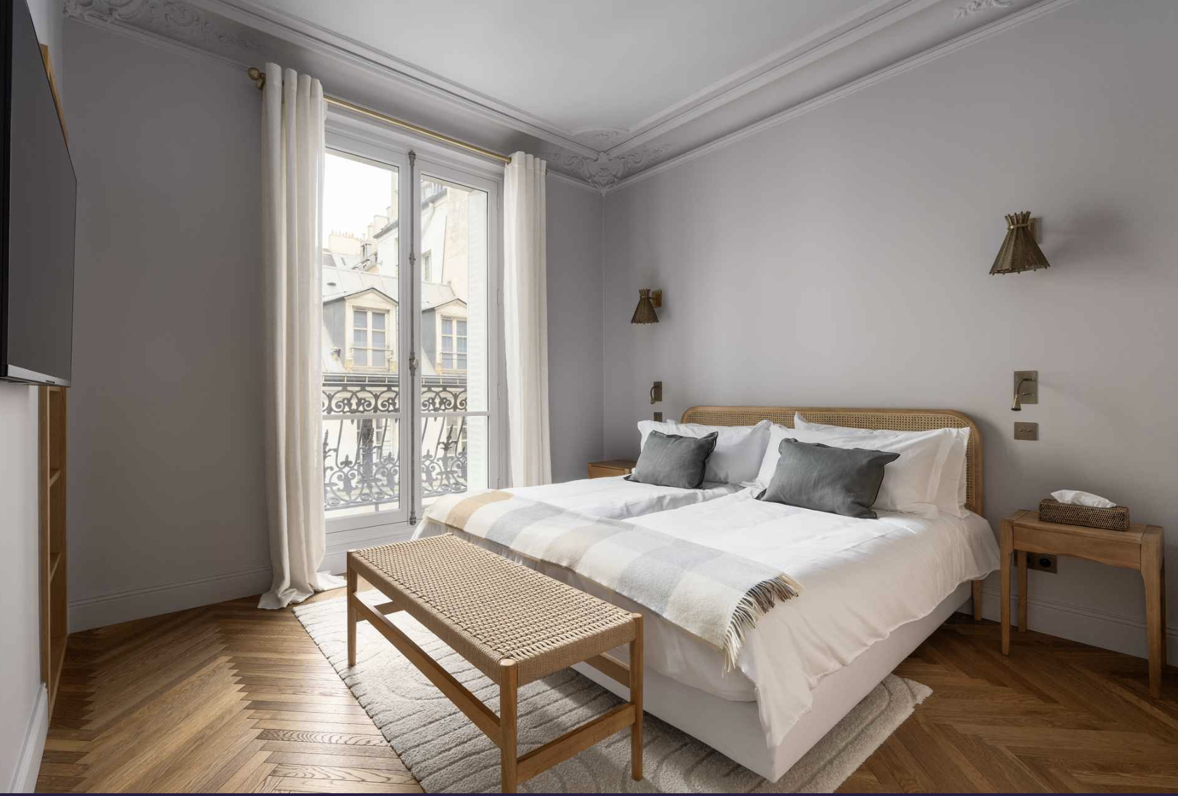 A bright hotel room with a large bed, gray pillows, and wooden furniture, including nightstands and a bench at the foot of the bed, with a window showing Parisian buildings outside.