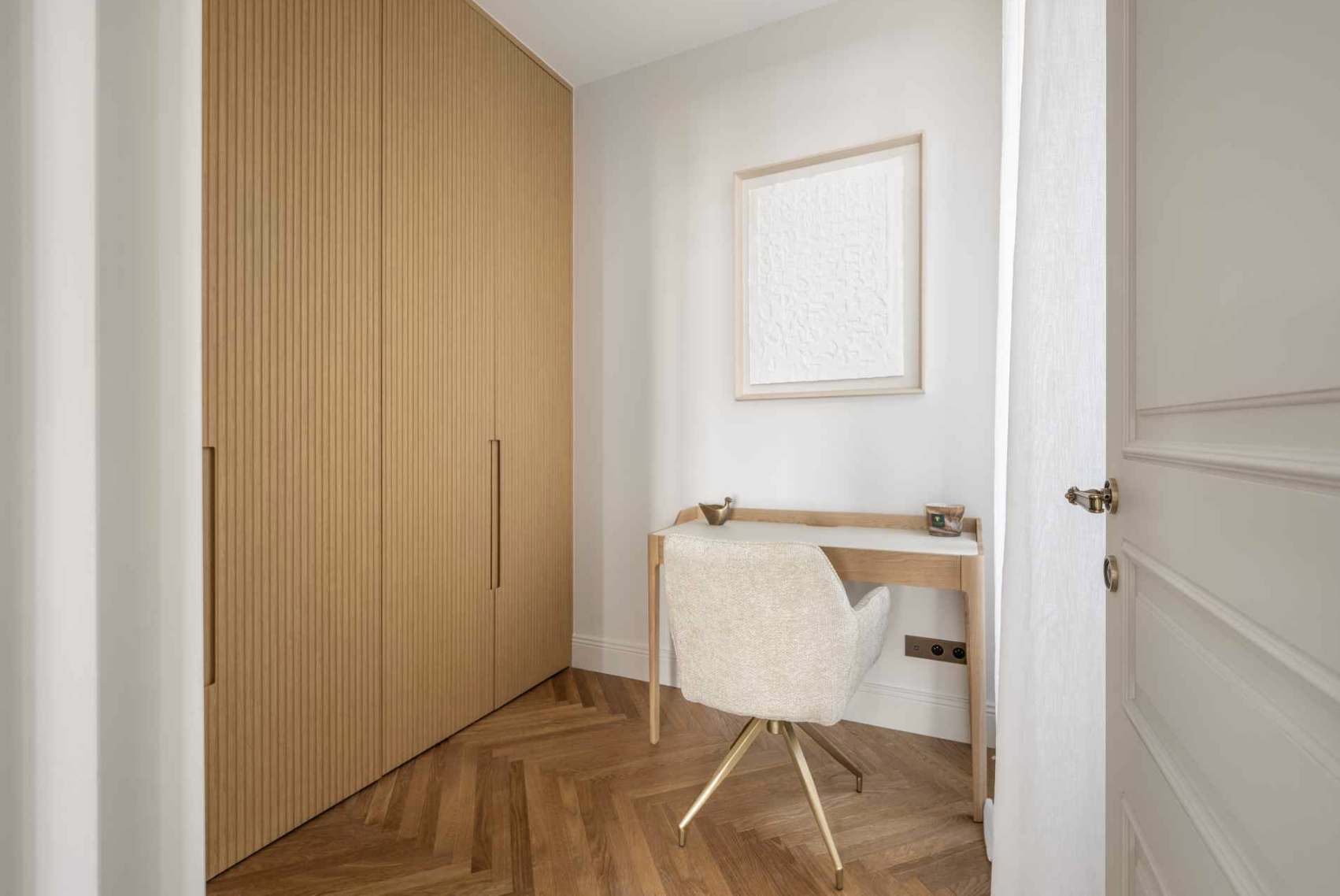 Small home office corner with a wooden desk, beige swivel chair, framed white textured wall art, and wooden floor.
