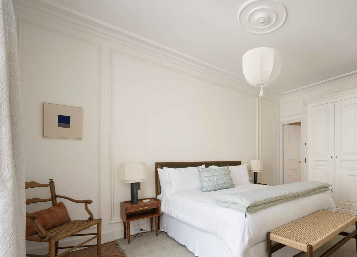 A bedroom with a white bed, bedside tables, lamps, a wooden bench at the foot of the bed, a wooden chair with a cushion, a closet with white doors, and minimalist artwork on the wall.