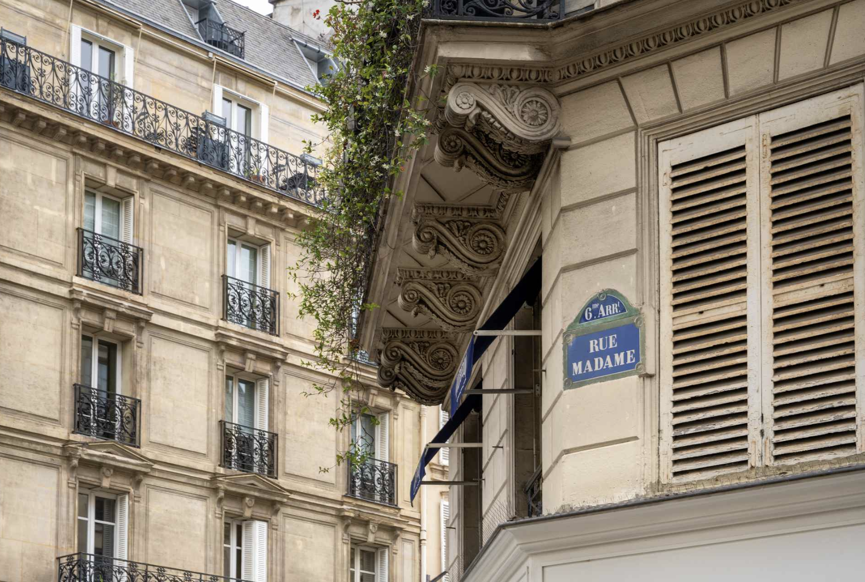Parisian building exterior with ornate architectural details, beige facade, window shutters, and black wrought iron balconies, with street sign for Rue Madame and 6th arrondissement.