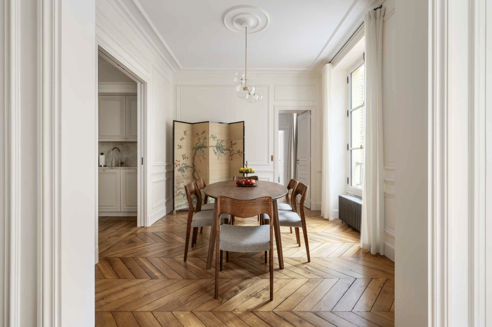 Dining room with a wooden table and six matching chairs, a decorative folding screen with floral design, a chandelier hanging from the ceiling, and large windows with white curtains, hardwood parquet flooring, and white paneled walls.
