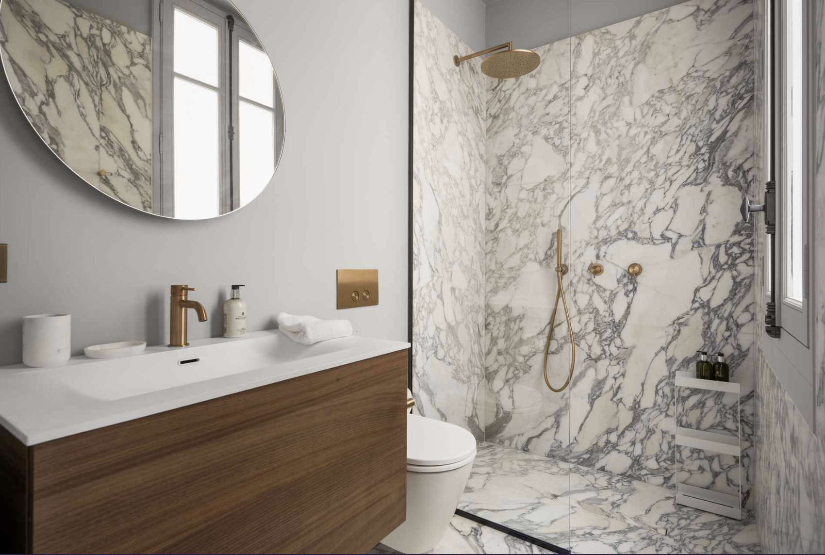 Modern bathroom with marble walls, a wooden vanity with a white countertop, a round mirror, a gold faucet, and a shower area with gold fixtures and a marble wall. There is a window letting in natural light.