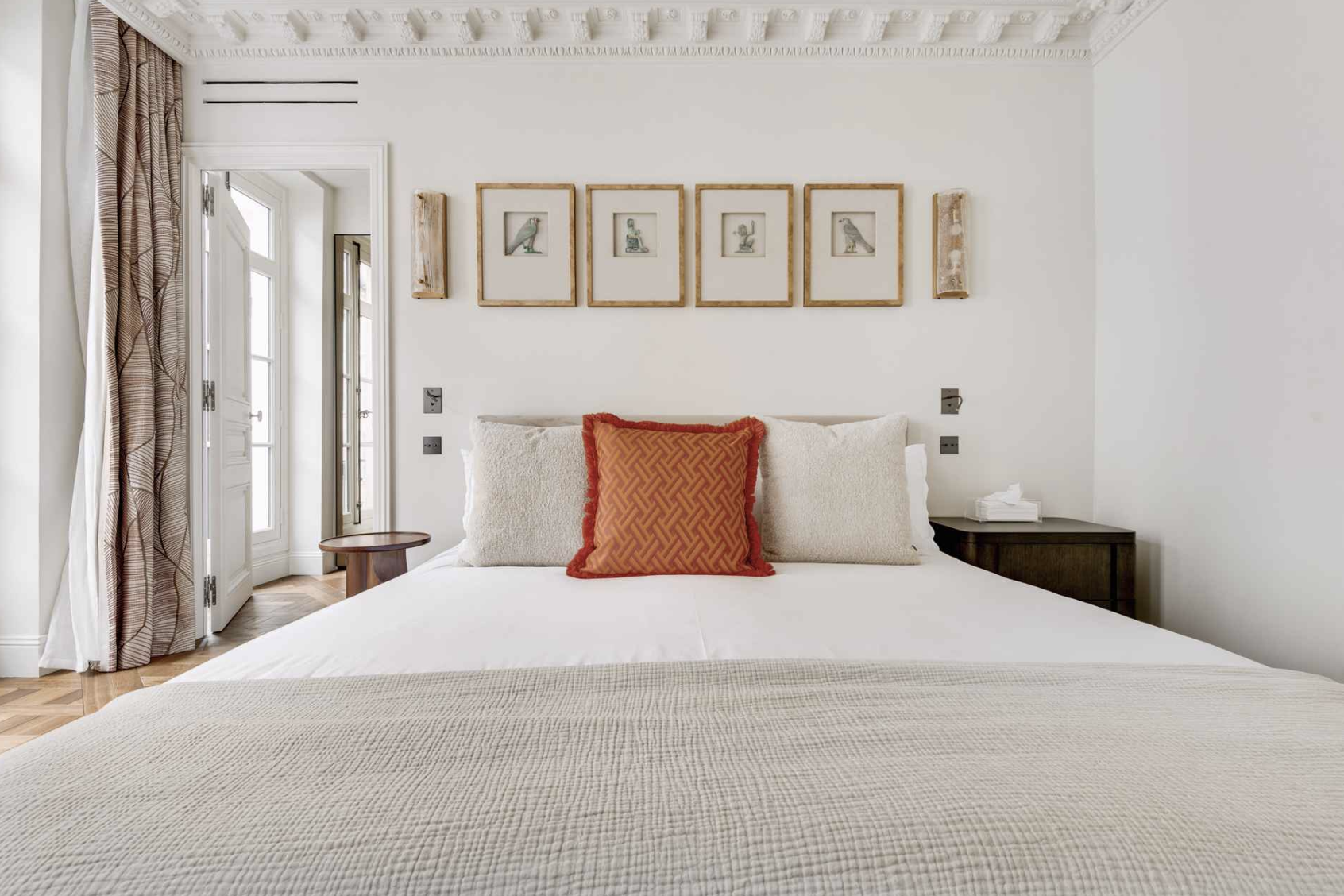 A neatly made bed with cream-colored pillows and a red decorative pillow in the center, in a bright, elegant bedroom with framed artwork of birds on the wall, a small bedside table with tissues, and a window with patterned curtains.