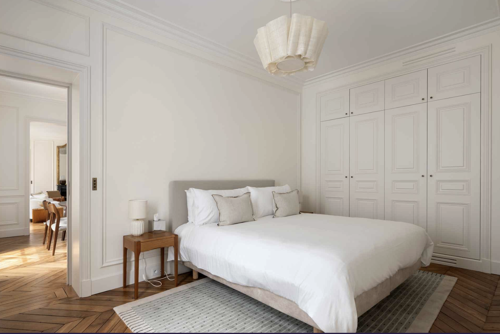 A bedroom featuring a bed with white bedding and multiple pillows, a small wooden nightstand with a white lamp, and built-in white closets. The room has white walls, molding, and a ceiling light fixture, with a wooden floor and an adjacent room visib
