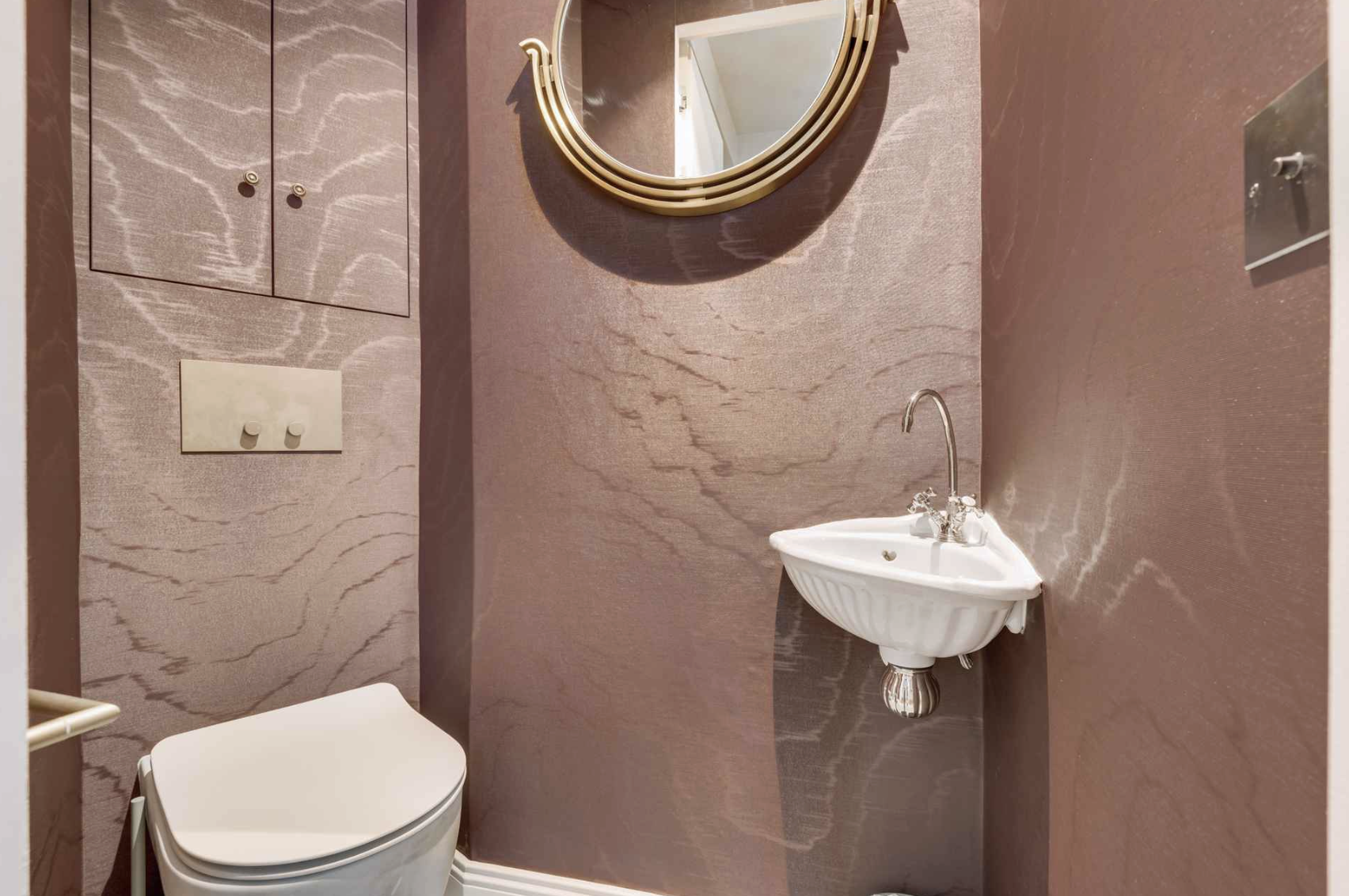 A small bathroom with pink marble walls, a white toilet, a small wall-mounted sink, a round mirror with a metal frame, and a wall-mounted light switch.
