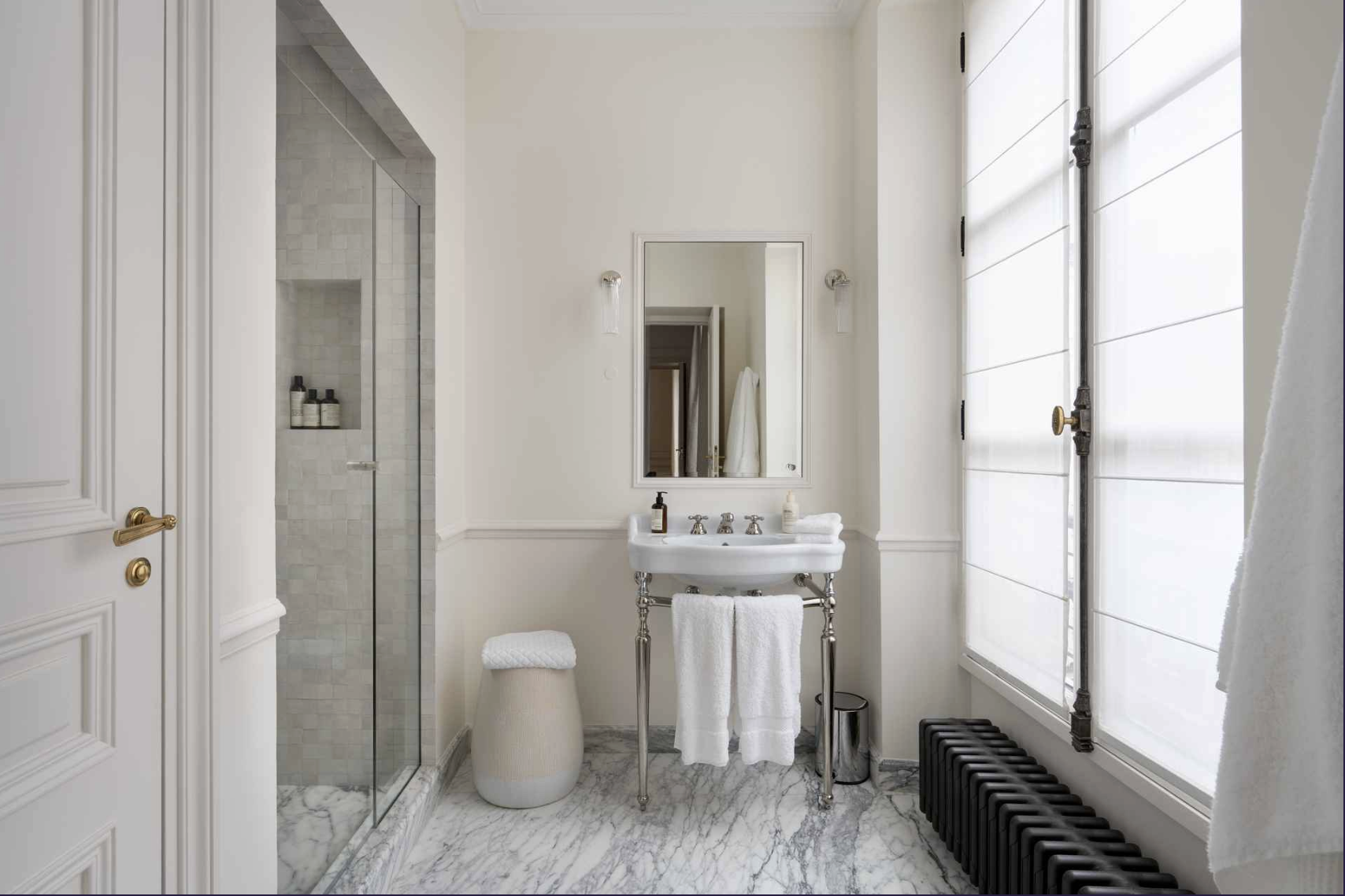 A modern, minimalist bathroom with a walk-in shower, small sink with silver legs, and a window with white blinds.