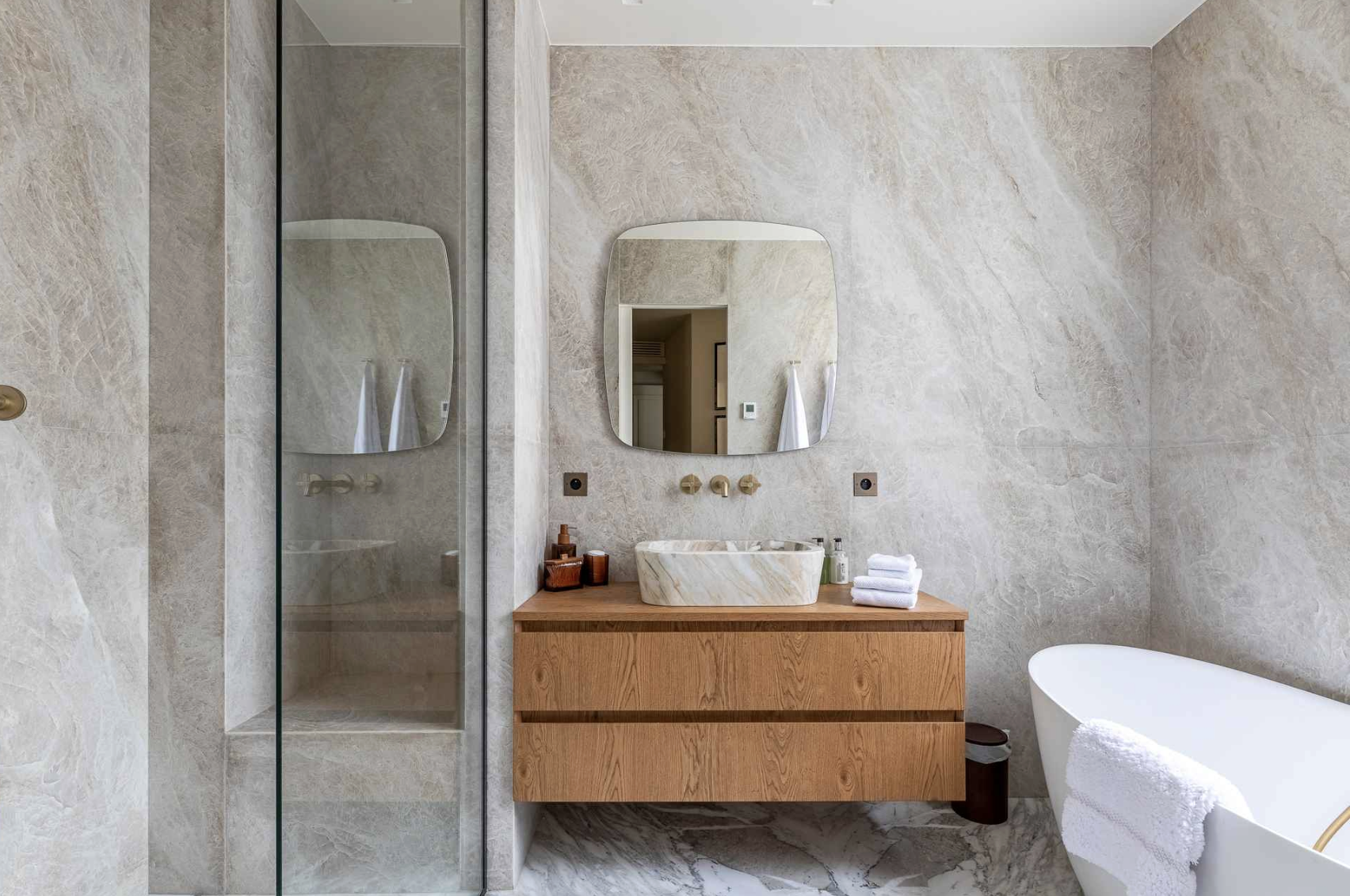 Modern bathroom with beige marble walls and floor, wooden vanity with marble sink, oval mirror, glass-enclosed shower, and a freestanding bathtub with a towel hanging over its edge.