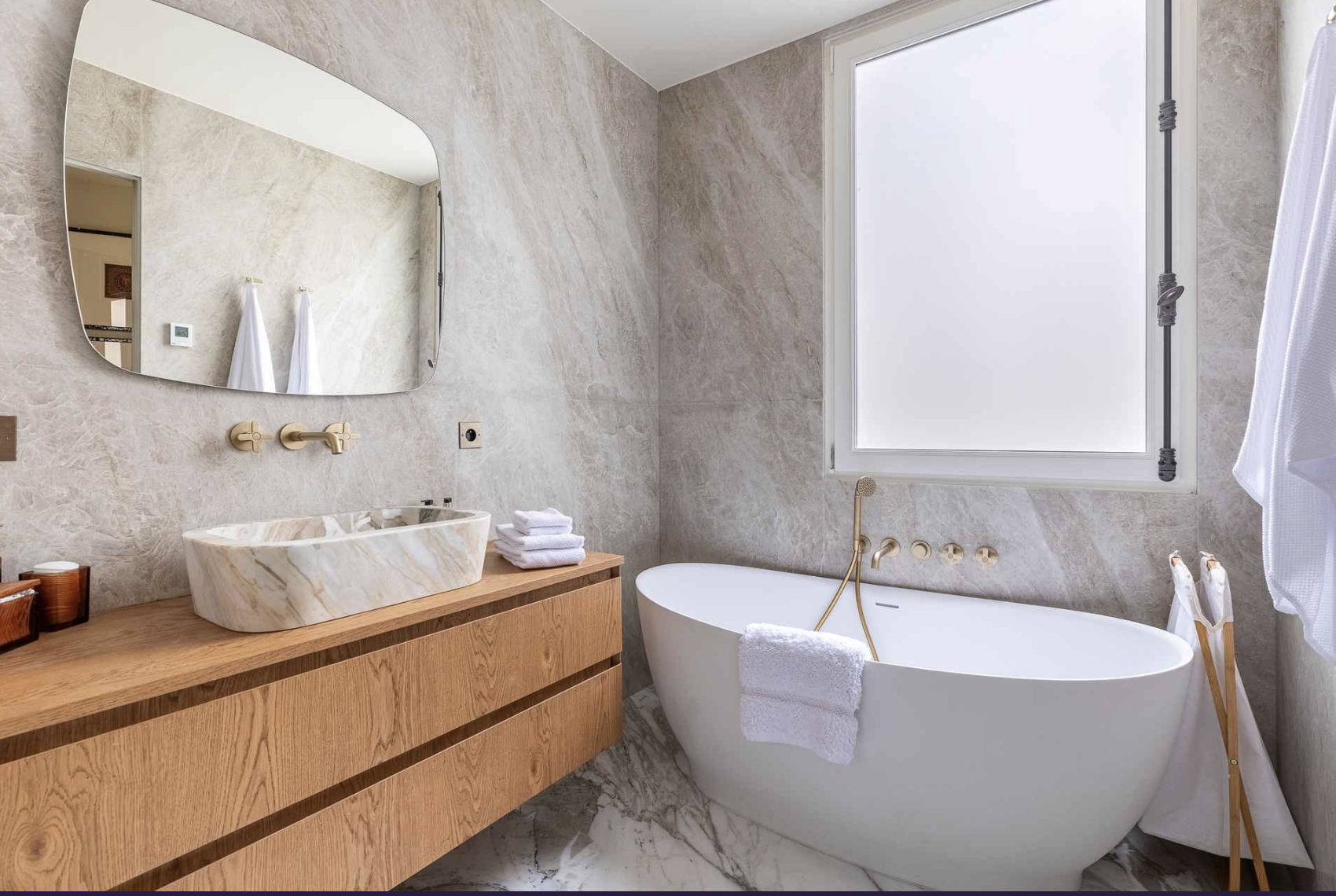 Modern bathroom with beige marble walls and floor, white bathtub, wooden vanity with marble sink, and large window with foggy glass