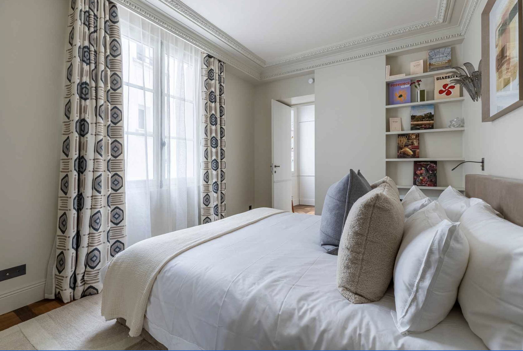 A neatly made bed with white linens and decorative pillows in a bright bedroom. There are patterned curtains, a window, and a white wall with a built-in shelf holding books and decorative items.