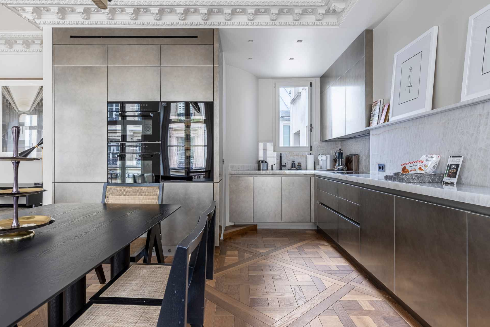 Modern kitchen with a black dining table, beige and gray cabinetry, hardwood parquet flooring, and a window above the sink.