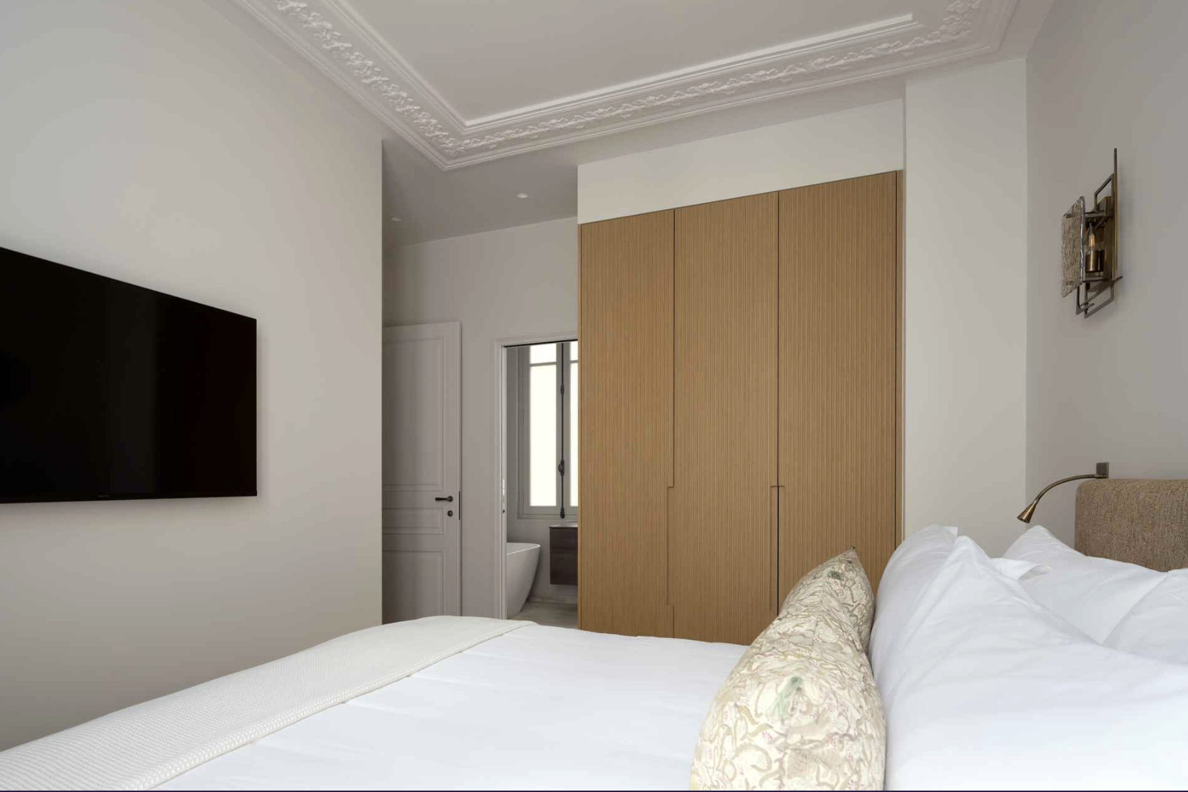 Modern bedroom with white walls, wooden closet, wall-mounted TV, bed with white linens and decorative pillow, small wall sconce, and a view of a bathroom through an open door.