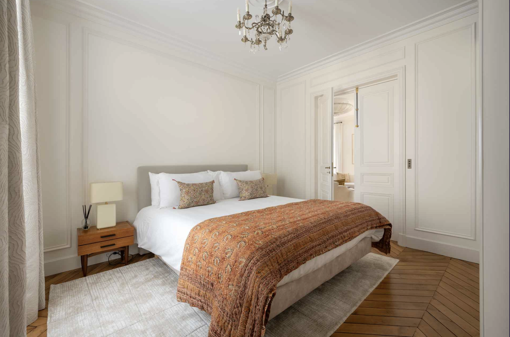 A bedroom with a large bed, white bedding, decorative pillows, a wooden nightstand, a lamp, a chandelier, and detailed white wall paneling.