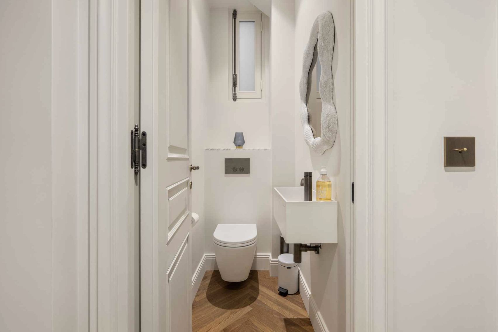 Small bathroom with a modern white toilet, a small white sink with a soap dispenser, a wavy mirror, a small window, and a brown wooden floor.