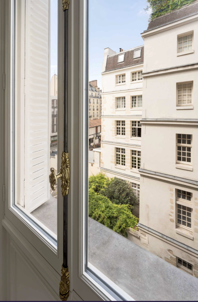 View through an open window showing neighboring beige buildings with multiple windows and a small outdoor area with green trees.