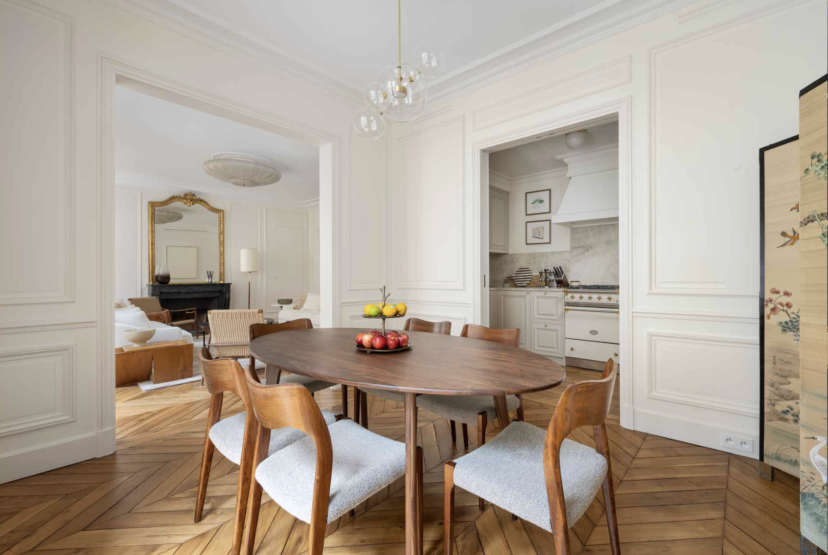 Dining room with wooden table and six chairs, fruits on the table, and an open view to living room and kitchen in a home interior.