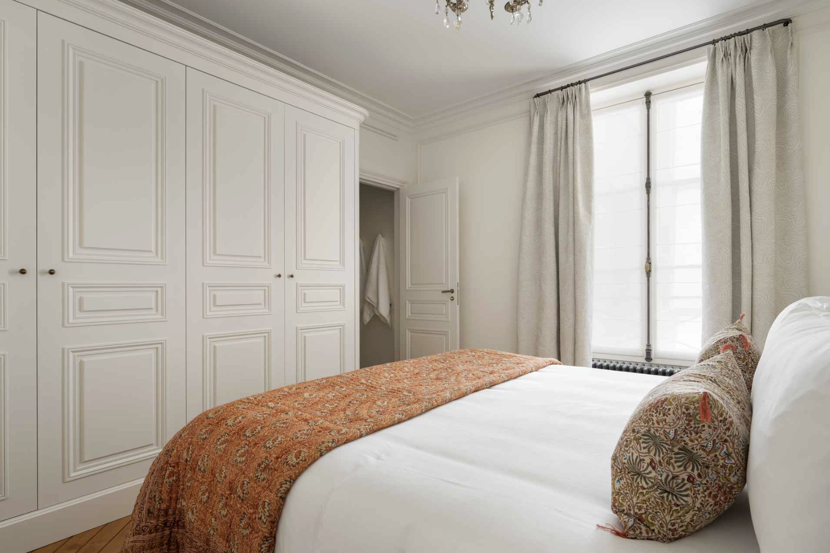 A bedroom featuring a bed with white sheets, patterned pillows, and a long orange-red blanket, next to a large window with light-colored curtains, white walls, and built-in closets.
