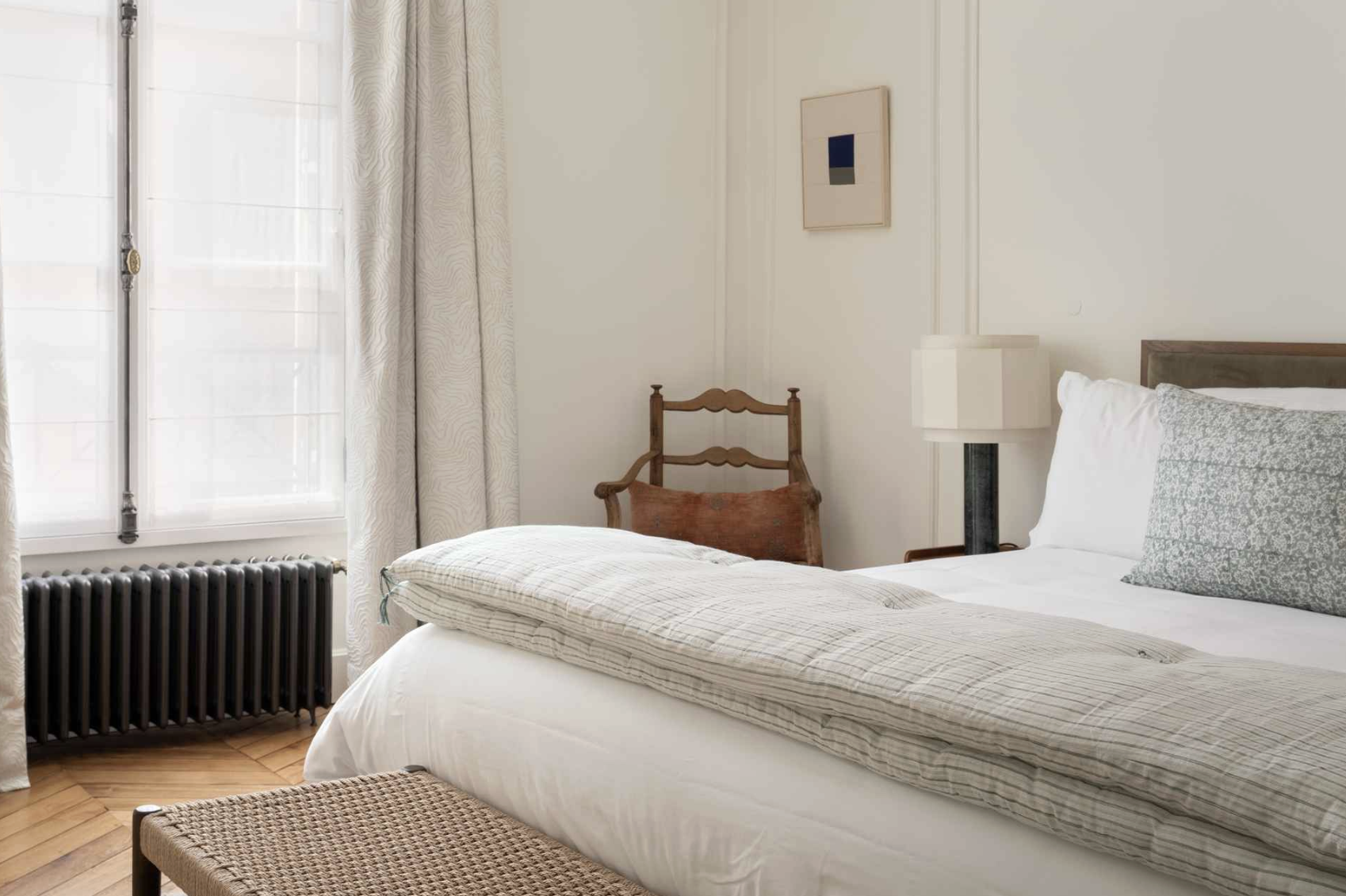 A bedroom with a bed, bedside lamp, framed artwork, a wooden chair, window with curtains, and a radiator.