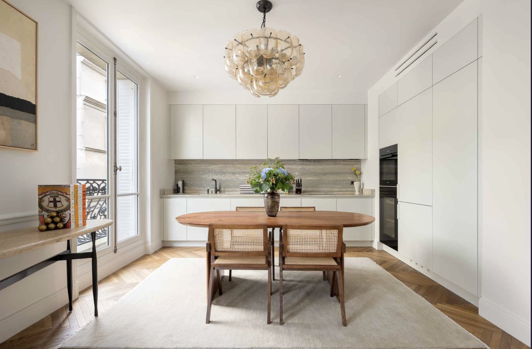 Bright modern kitchen with white cabinets, a wooden dining table with two chairs, a vase of flowers, and large windows with natural light.