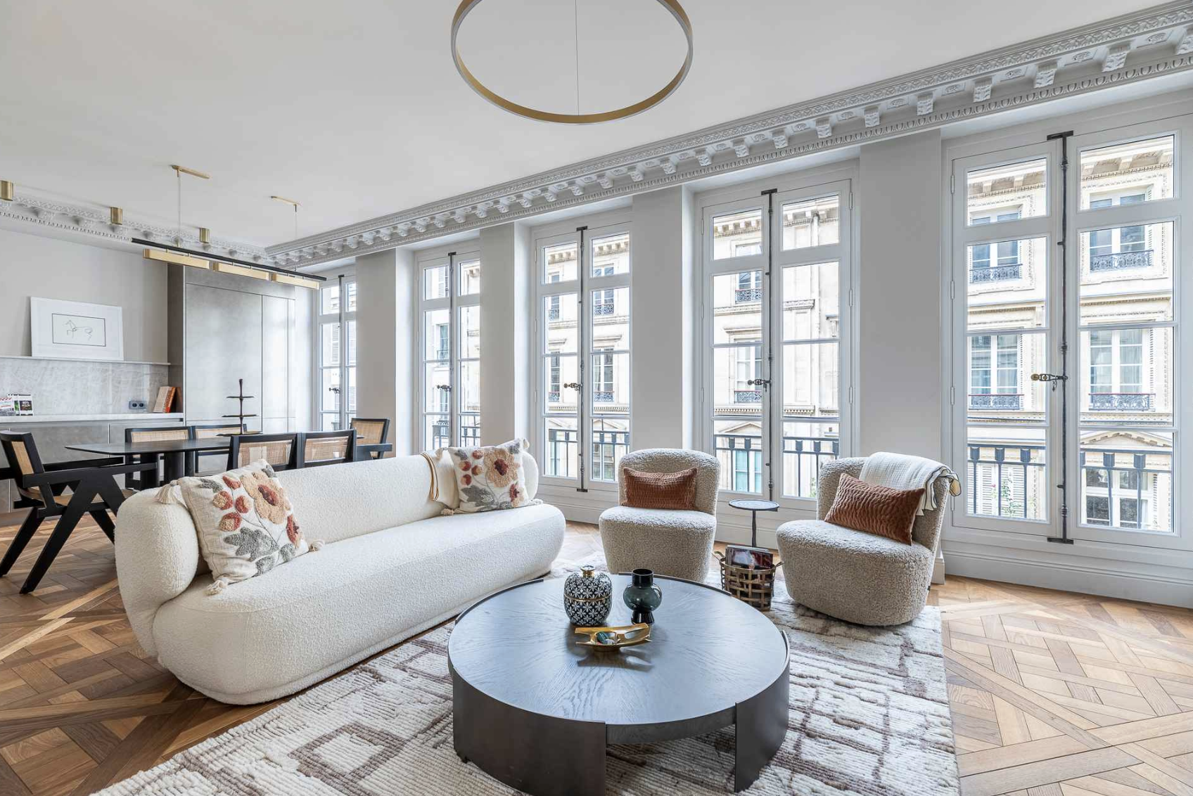 Bright living room with large windows, white walls, a white sofa with floral pillows, two matching armchairs with rust-colored pillows, a round black coffee table with decorative items, and a dining area with a black table and chairs in the backgroun