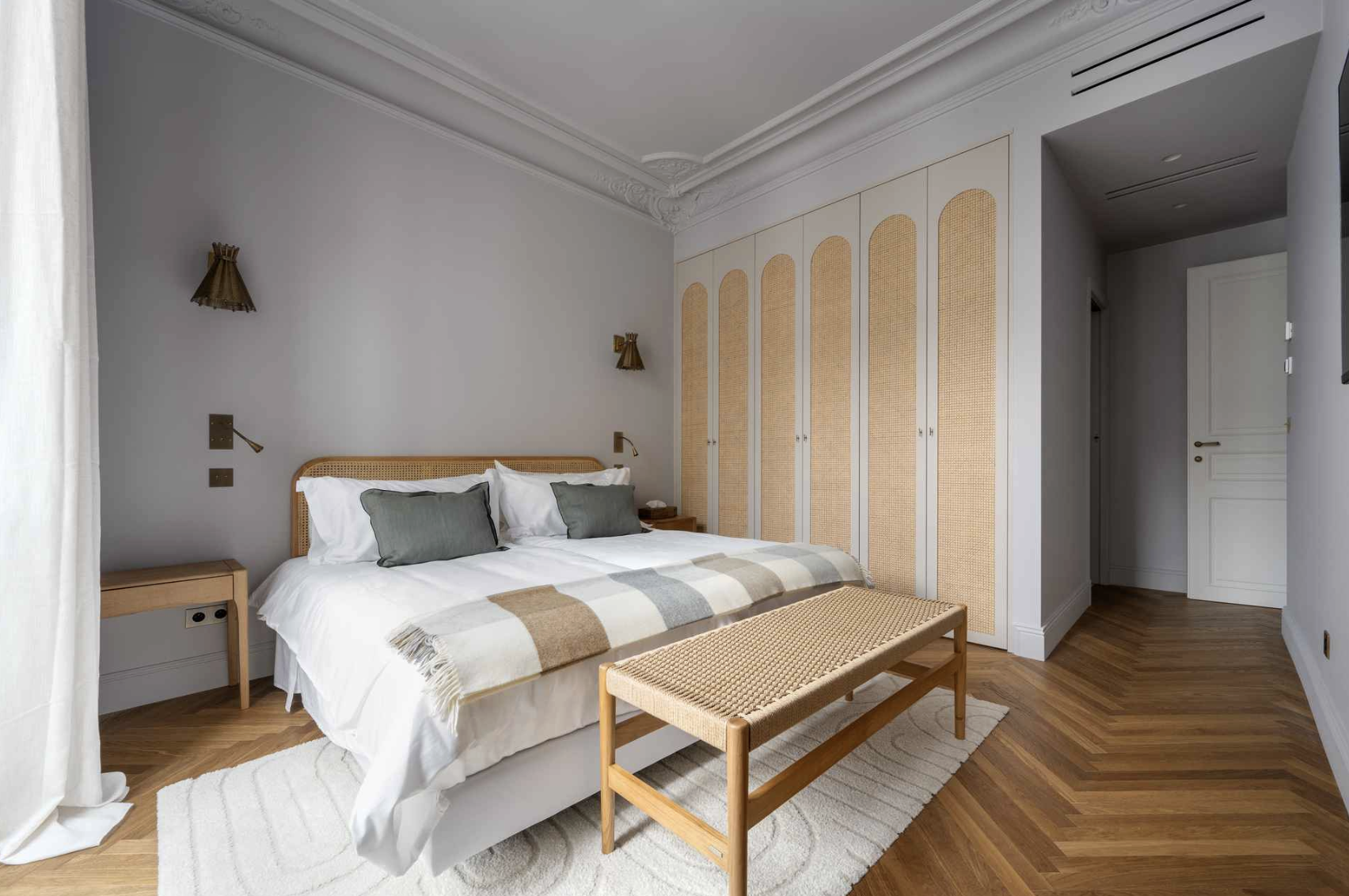 Modern bedroom with a bed, gray pillows, beige and white blanket, wooden bench, nightstands, large corner closet, hardwood floors, and gray walls.