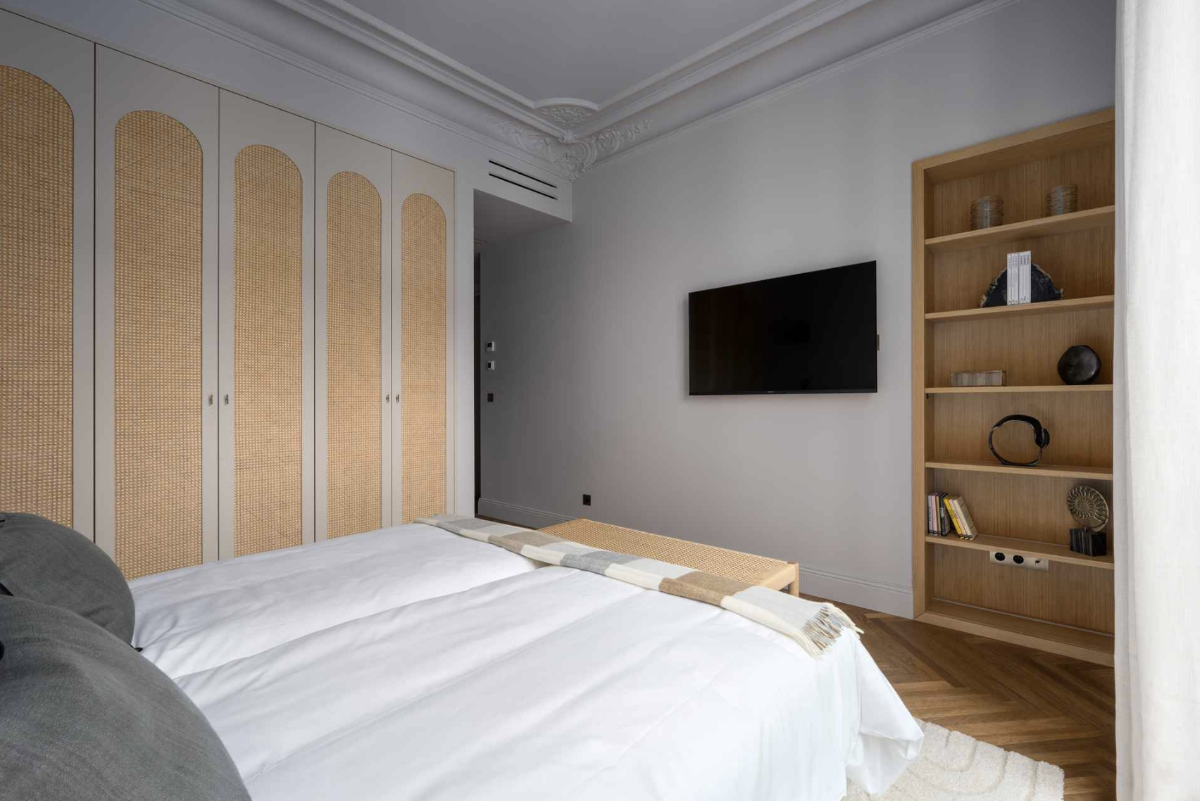 Bedroom with a bed, a built-in wardrobe with cane-front doors, a wall-mounted TV, and a wooden bookshelf with decorative items and books.