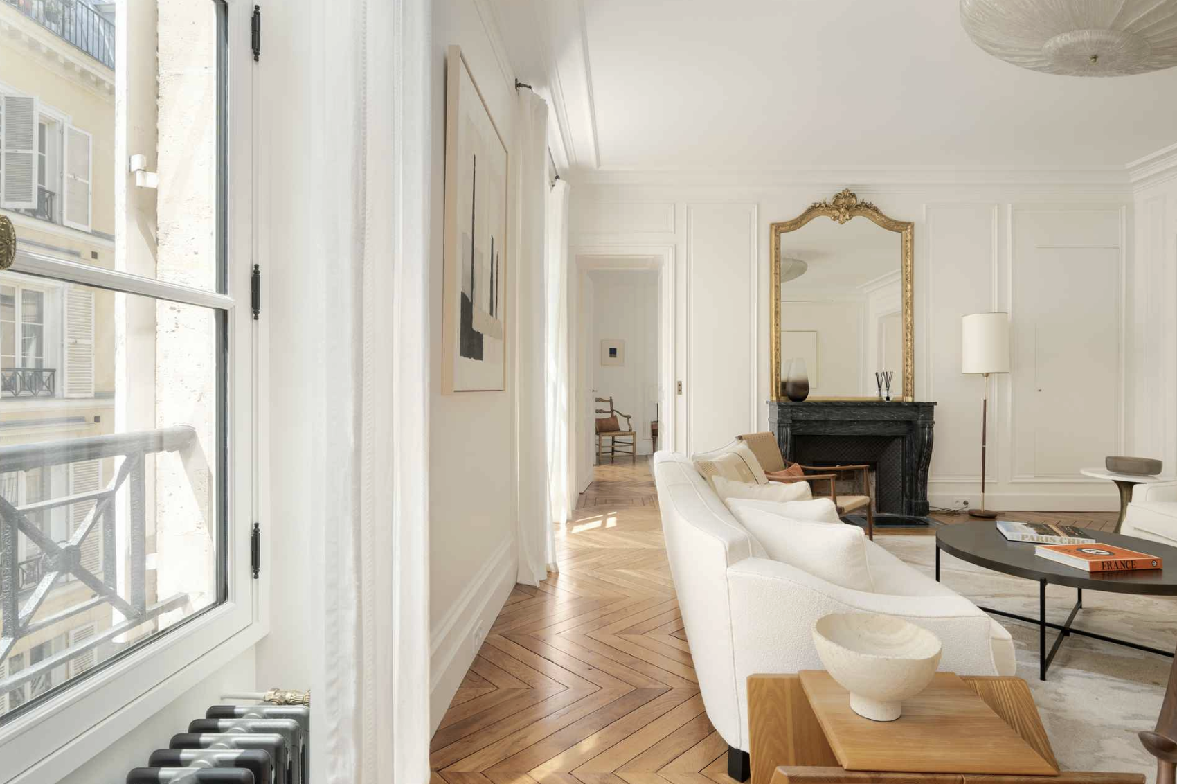 Bright living room with white walls, hardwood herringbone floors, large window, white sofa, black fireplace with gold mirror above, tall floor lamp, small side table, coffee table with books, and art on the walls.