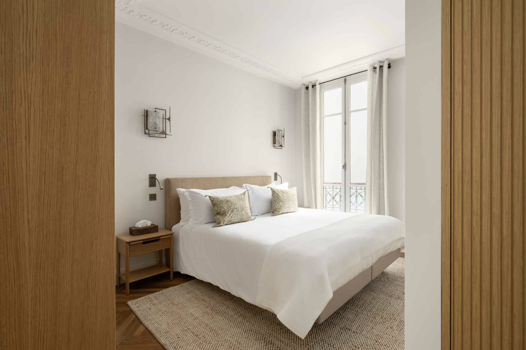 A modern bedroom with a white bed, beige headboard, nightstand, and decorative pillows; large window with sheer curtains and iron balcony railing visible outside.