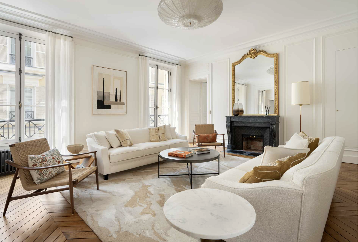 Living room with white sofas, wooden chairs, a black fireplace, large windows with white curtains, a mirror above the fireplace, and artwork on the wall.