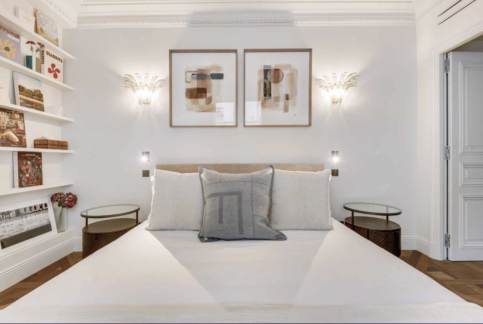 A bedroom with a bed, three pillows, two wall sconces, two abstract art pieces above the bed, and two bedside tables with glass tops.