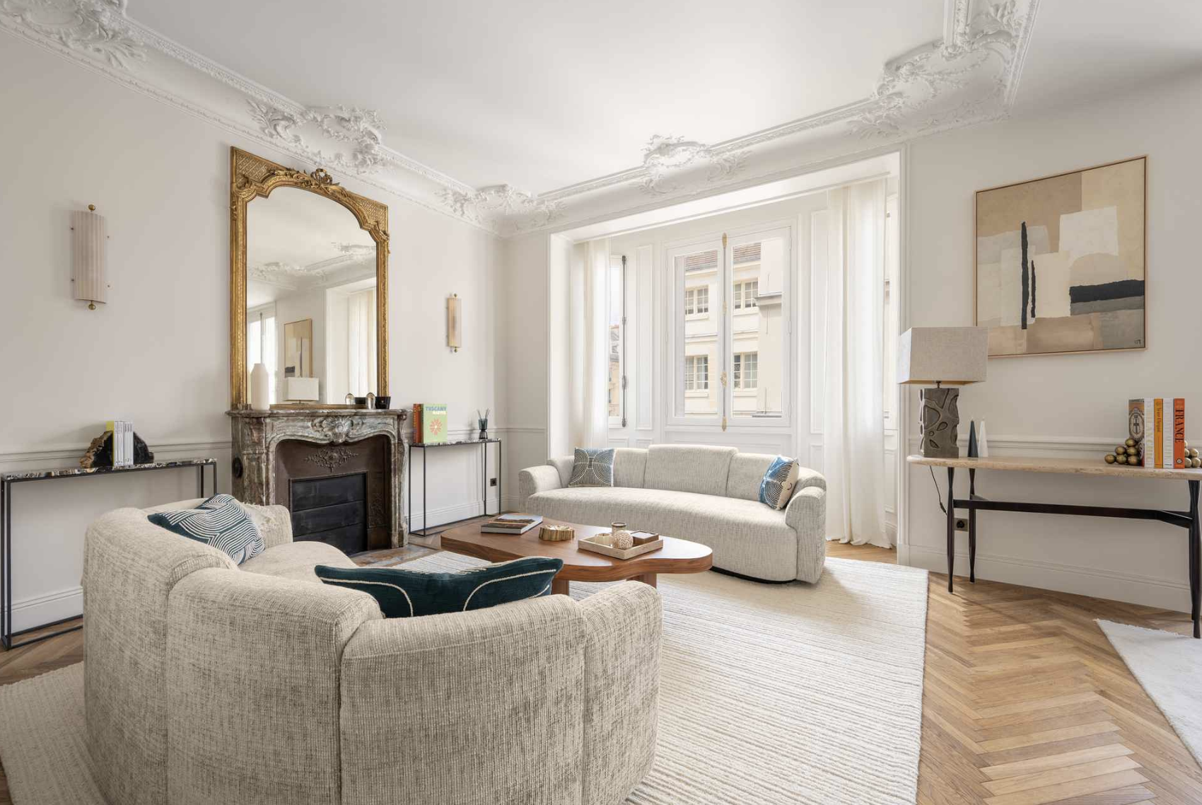 Living room with white walls, ornate ceiling molding, large window with curtains, cream sofa, chairs, wooden coffee table, marble fireplace with a gold mirror above, abstract wall art, side tables with books and decor, floor lamp, light-colored rug,