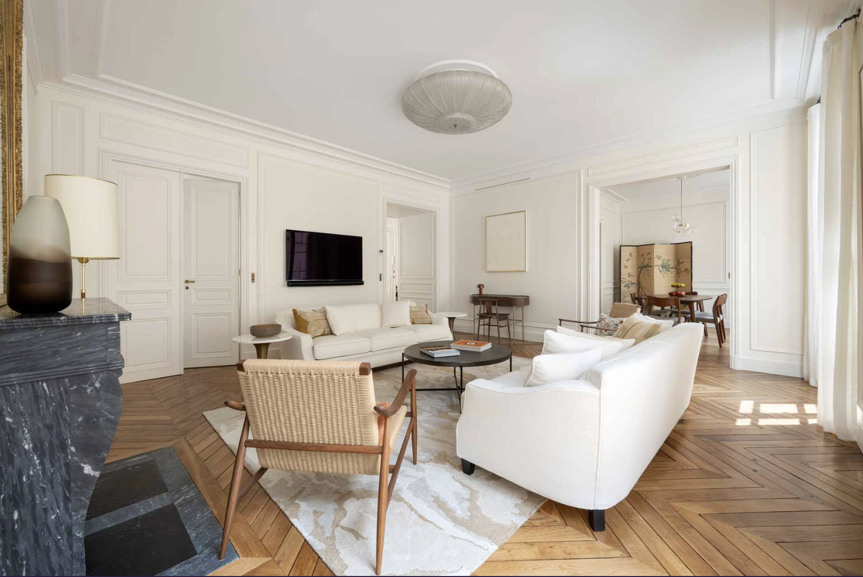Living room with white walls, hardwood floors, a white sofa, an armchair, a round black coffee table, a wall-mounted TV, and a dining area with a table and chairs in the background.
