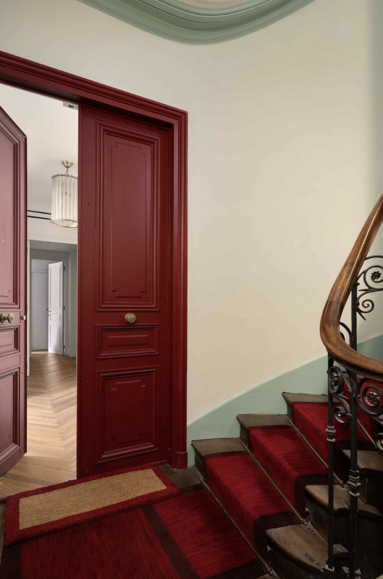 Interior view of a house featuring a staircase with red carpet and ornate black handrail, a red door with decorative panels leading to another room, a mat at the base of the stairs, and a view of a room with light-colored walls, a chandelier, and woo